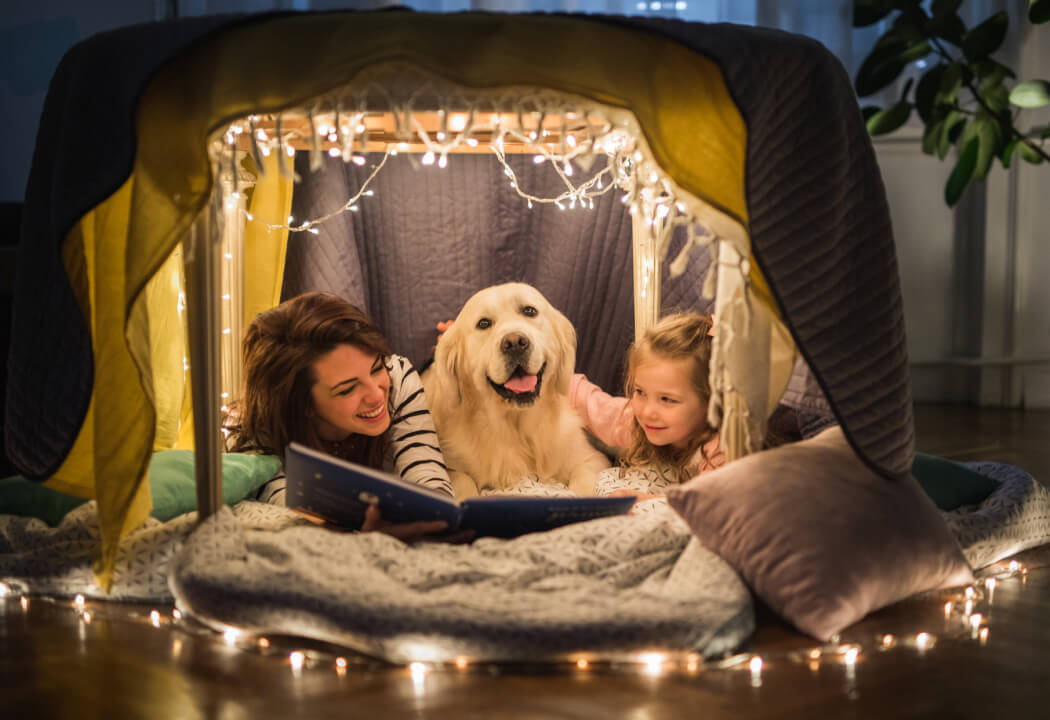 Mom, young girl, and dog huddled under blankets and reading a story book 
